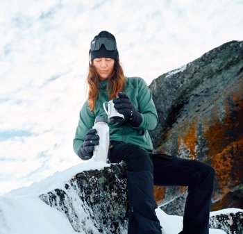 Isolierte silberne Wasserflasche und schwarzer Becher, gehalten von einer Person in einer grünen Fleecejacke und schwarzen Handschuhen, die auf einem verschneiten Felsen sitzt.