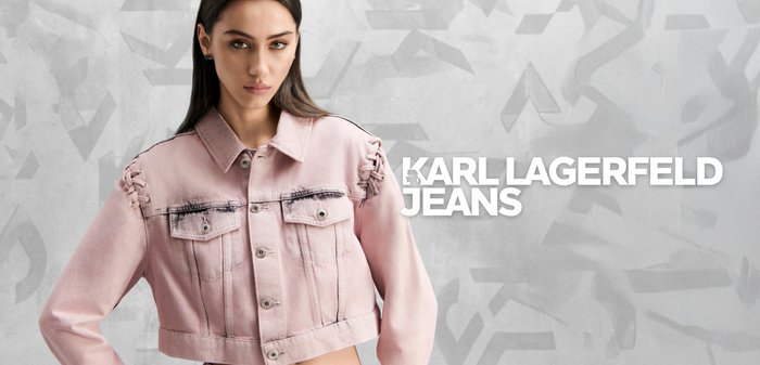 Femeie purtând o jachetă scurtă din denim roz deschis și blugi asortați, stând cu o mână pe șold, pe un fundal gri cu model.