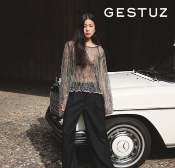 Prozirni top s dugim rukavima ukrašen perlicama, s zakrivljenim rubom, uparen s crnim širokim hlačama. Iza se vidi elegantan vintage automobil.