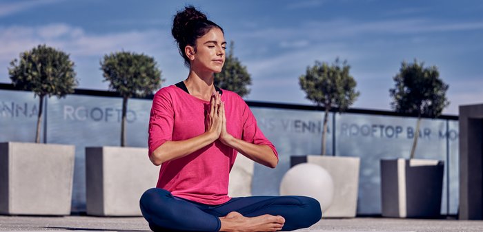 Roze top met lange mouwen, zwarte tanktop eronder en marineblauwe leggings. Vrouw in yogahouding, handen samengebracht, zittend op een glad oppervlak met een stadsachtergrond.