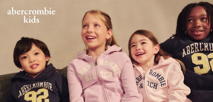 Trei copii zâmbitori purtând hanorace Abercrombie Kids, stând aproape unul de altul, pe un fundal simplu, cu numele brandului deasupra.
