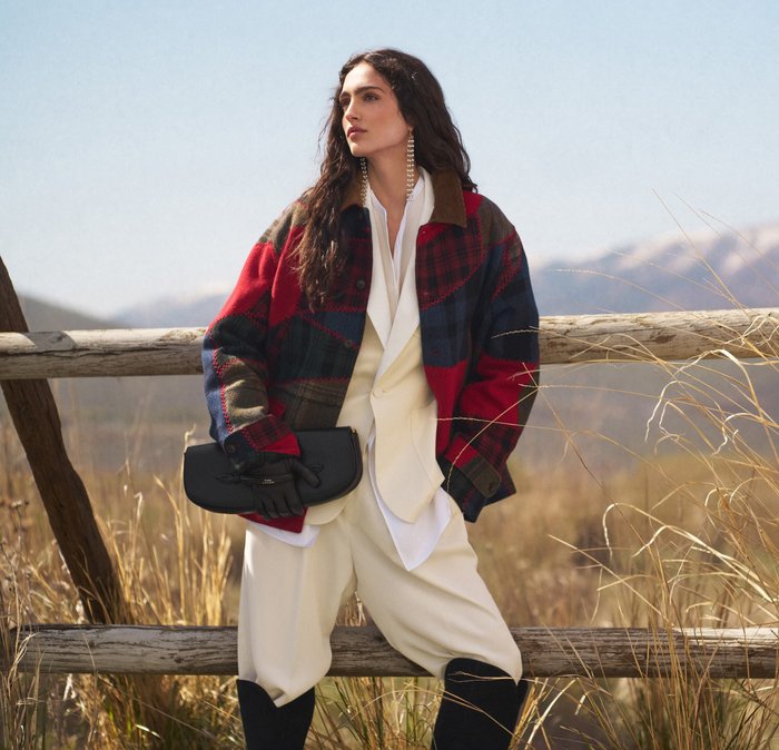 A woman in a cream suit and red plaid jacket leans against a wooden fence in tall grass, holding a black clutch and wearing gloves, with mountains in the background.