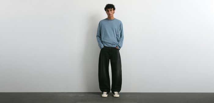 Jeune homme debout contre un mur blanc, portant une chemise à rayures bleues à manches longues, un pantalon large noir et des baskets blanches avec des détails noirs.