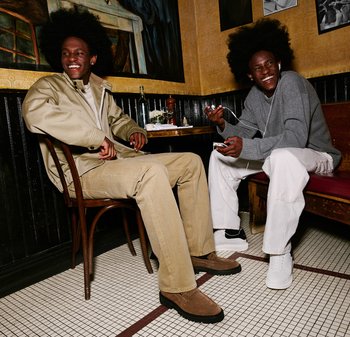 Twee personen zitten in een restaurant, gekleed in casual outfits: de een in een beige jas en broek met bruine schoenen, de ander in een grijze trui en witte broek.