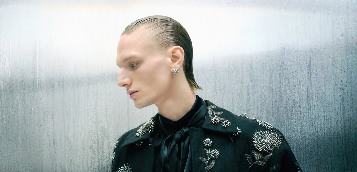 Abrigo negro bordado con diseños florales, corbata de seda y arete de plata. El sujeto tiene el cabello peinado hacia atrás contra un fondo brumoso.