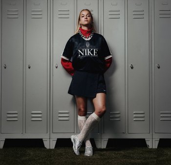 Schwarzes Nike-Trikot mit weißen Akzenten, rotem Kragen und einem schwarzen Rock. Weiße Spitzenkniestrümpfe und silberne Sportschuhe. Graue Spinde im Hintergrund.