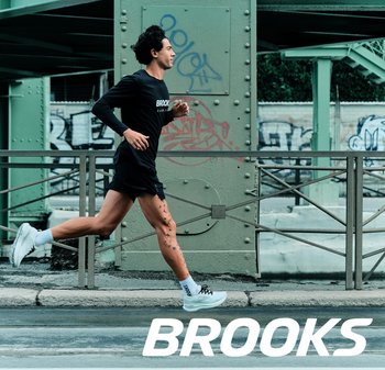 Man met tatoeages die rent langs een stedelijke straat onder een groene metalen brug, gekleed in een zwart hardloopoutfit van Brooks en witte Brooks schoenen.
