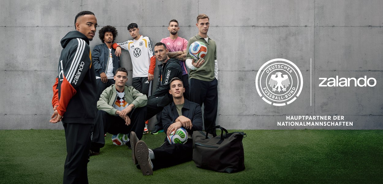 Acht Männer posieren in Sportkleidung auf Kunstrasen mit Fußbällen, einem Sporttasche und einem Hintergrund aus Betonwänden mit den Logos des Deutschen Fußball-Bunds und Zalando.