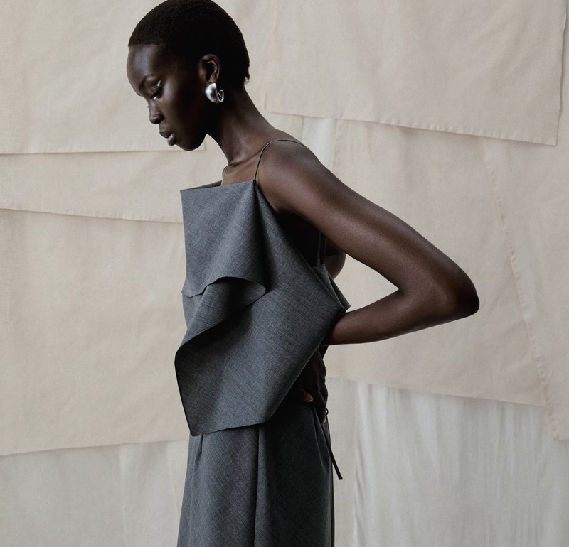 Femme aux cheveux courts portant une robe grise asymétrique et de grandes boucles d'oreilles en argent, posant de profil devant un fond de tissu beige.