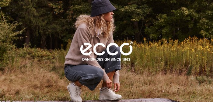 Femme aux cheveux bouclés, vêtue de vêtements décontractés et d'un chapeau seau, accroupie en plein air, attachant ses bottes blanches sur une bûche dans un cadre naturel.