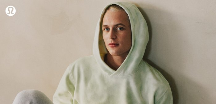 Una giovane persona con una felpa con cappuccio verde chiaro indossata con il cappuccio alzato, seduta contro un muro beige con un'espressione neutra.