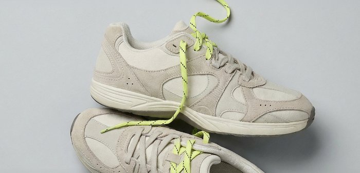 Beige Wildleder-Sneaker mit weißer Sohle, ausgestattet mit doppelten Schnürsenkeln – einem grauen und einem gelben mit schwarzen Streifen, nebeneinander gebunden.