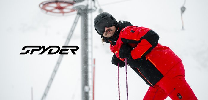 Veste de ski rouge et noire avec des accents orange, col montant, pantalon assorti, flocons de neige visibles, lunettes de ski portées, télésiège en arrière-plan.