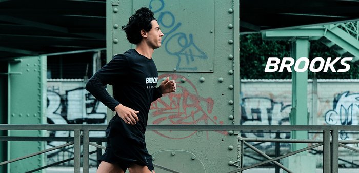 Man in zwarte Brooks hardloopkleding rent voorbij een groene metalen brug met graffiti in een stedelijke omgeving.
