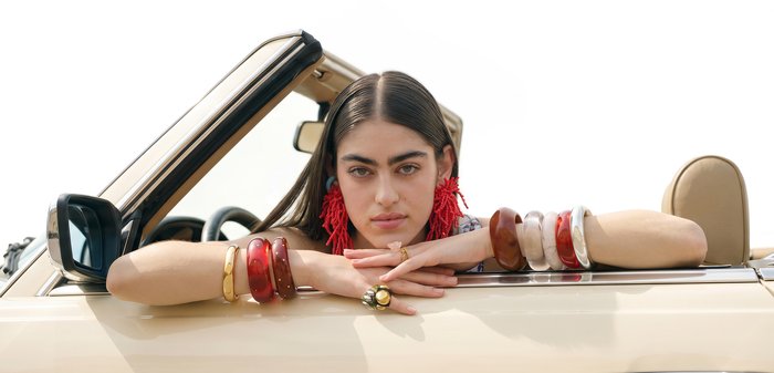 Femme aux longs cheveux foncés, boucles d'oreilles rouges audacieuses et plusieurs bracelets, reposant ses bras sur la porte d'une voiture décapotable beige.