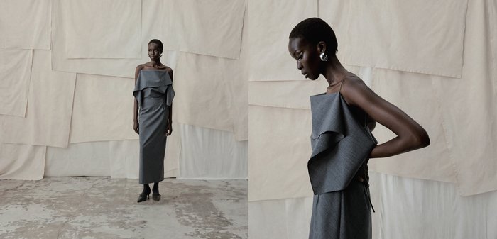 Γυναίκα με γκρι, ασύμμετρο φόρεμα έξω από τον ώμο με λεπτομέρειες βολάν, στέκεται μπροστά σε ένα υφασμάτινο, ουδέτερου τόνου φόντο.