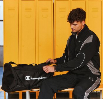 Black duffel bag with white "Champion" logo, smooth fabric, curved shape, and dual handles, placed on a bench near yellow lockers.