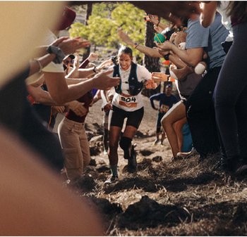 Coureur en chemise blanche et shorts noirs, portant un gilet d'hydratation, naviguant sur un sentier rocailleux avec une foule encourageante de chaque côté.