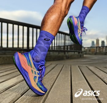 Loper met paarse en oranje ASICS-schoenen en paarse sokken, zwevend in de lucht op een houten loopbrug met de skyline van de stad op de achtergrond.