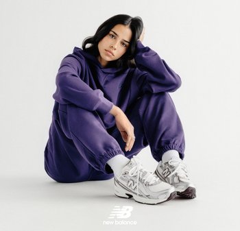 Mujer con cabello negro sentada en el suelo vistiendo una sudadera con capucha morada, pantalones deportivos a juego, zapatillas blancas New Balance y calcetines blancos sobre un fondo blanco.