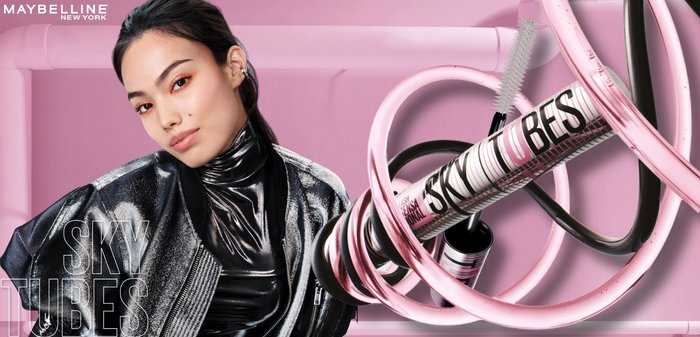 Woman with glossy black outfit and orange eye makeup next to Maybelline Sky Tubes mascara with swirling pink and black tubes on a pink background.