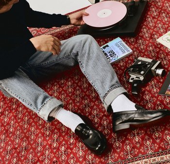 Mocassins en cuir noir avec une finition lisse, accompagnés de chaussettes blanches côtelées et d'un jean bleu clair, posés sur un tapis rouge à motif.