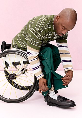 Mann im Rollstuhl passt den unteren Teil seiner grünen Hose über seinem schwarzen Anzugschuh vor einem blassrosa Hintergrund an.