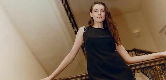 Kvinna med långt brunt hår, klädd i en ärmlös svart klänning, lutar sig mot en träbalustrad inne i en byggnad med en trappa i bakgrunden.