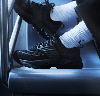 Personne portant des chaussures de sport noires et des chaussettes blanches côtelées avec un logo noir, assise avec les pieds posés sur une surface métallique.