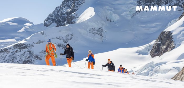 Gruppe von Bergsteigern, die zusammen gesichert über eine verschneite Berglandschaft unter klarem Himmel trekkt.