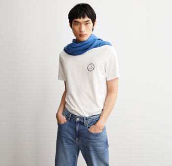 Weisses Baumwoll-T-Shirt mit einem runden schwarzen Smiley-Gesicht-Logo, kombiniert mit einem blauen Stoff um den Hals, getragen mit blauen Jeans.