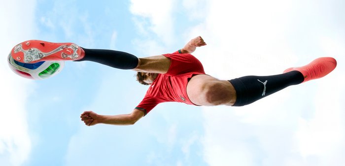 Παίκτης ποδοσφαίρου με κόκκινη φανέλα και μαύρες κάλτσες εκτελεί ψηλό κτύπημα με φόντο τον γαλάζιο ουρανό.