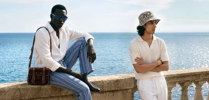 Twee stijlvolle mannen poseren bij een stenen balustrade met de zee achter hen; de een zit met een zonnebril en gestreepte broek, de ander staat in het wit met een hoed met patroon.