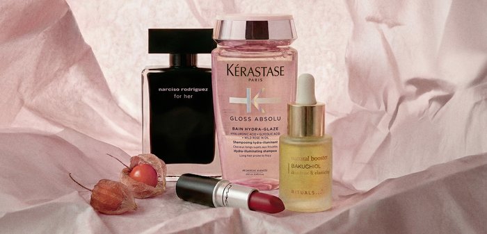 Parfum Narciso Rodriguez, shampooing Kérastase, sérum Bakuchiol et rouge à lèvres MAC rouge disposés sur un tissu rose avec des fruits physalis séchés.