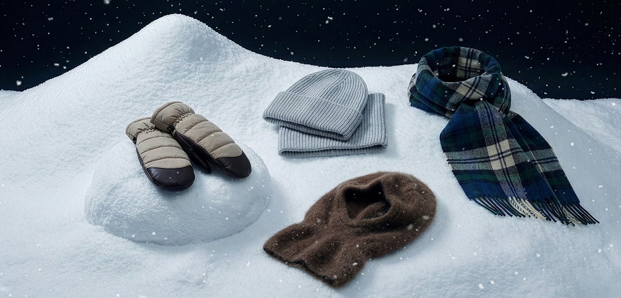 Polstrede beige votter med brune detaljer, grå strikkelue, lys grå skjerf, mørk brun strikkelue og rutete skjerf på snø.