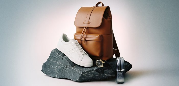 Sac à dos en cuir marron, basket blanche, montre-bracelet noire avec détails dorés, et flacon de soin de la peau sur une surface en pierre texturée foncée.