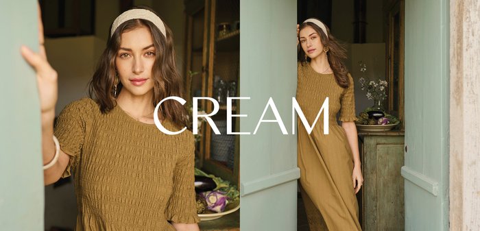 Femeie într-o rochie galben muștar, stând în cadrul ușii unei bucătării rustice, lângă un dulap cu un bol de legume și un vas cu flori.