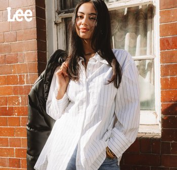 Camicia bianca a righe con vestibilità comoda, design con bottoni, abbinata a jeans in denim. Il modello tiene una giacca a vento nera appoggiata sulla spalla.