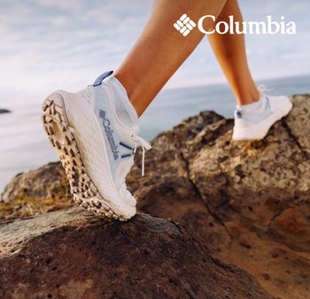 Des chaussures Columbia blanches avec des accents bleus, des semelles en caoutchouc texturé et un dessus en maille, positionnées sur un terrain rocheux contre un fond doux.