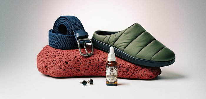 Vihreä toppattu liukuselkine, sininen kudottu vyö, partaöljypullo, jossa etiketti "Sitrus", ja pyöreät kalvosinnapit aseteltuna punaisen karkeapintaisen kiven päälle.