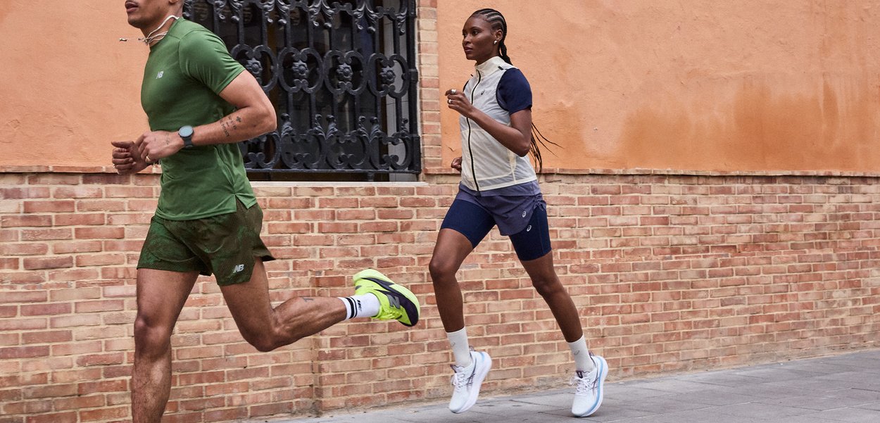 Άντρας και γυναίκα τρέχουν έξω δίπλα σε τοίχο από τούβλα και παράθυρο, φοράνε αθλητική ένδυση και αθλητικά παπούτσια.