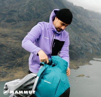 Lila Fleecejacke mit schwarzen Akzenten, getragen von einer Person, die eine türkis- und grau farbige Outdoorjacke mit Kapuze auspackt.