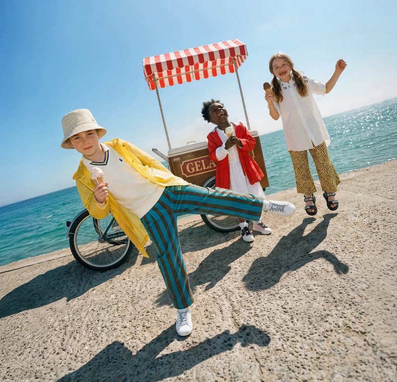 Tres niños comiendo helado junto a un carrito de gelato junto al mar, con un niño levantando una pierna juguetonamente y los demás sonriendo y saltando.