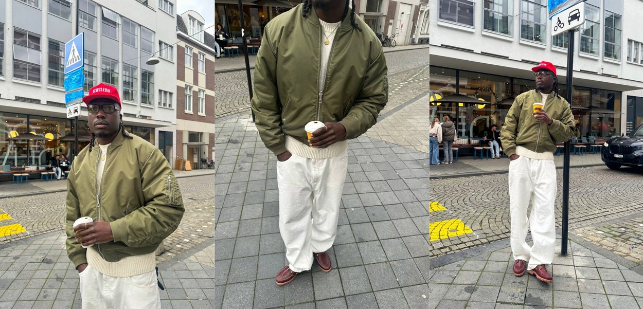 Man met een groene bomberjack, rode pet, witte broek en bruine schoenen die een koffiekop vasthoudt op een stedelijke straat met winkels en een zebrapad.