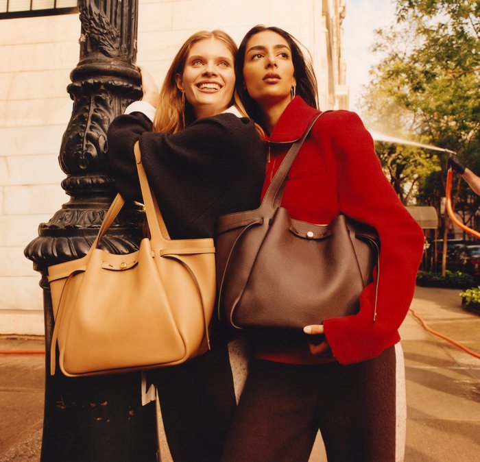 Due donne posano insieme mentre tengono grandi borse a tracolla in pelle. Una borsa è color beige con un accento dorato; l'altra è marrone scuro con pelle testurizzata.