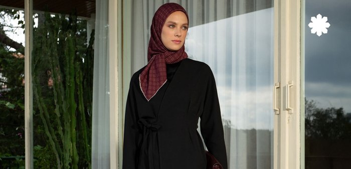 Veste noire à nouer à la taille, associée à un hijab à motifs bordeaux et une jupe lisse crème. Tiens un sac à main bordeaux foncé.