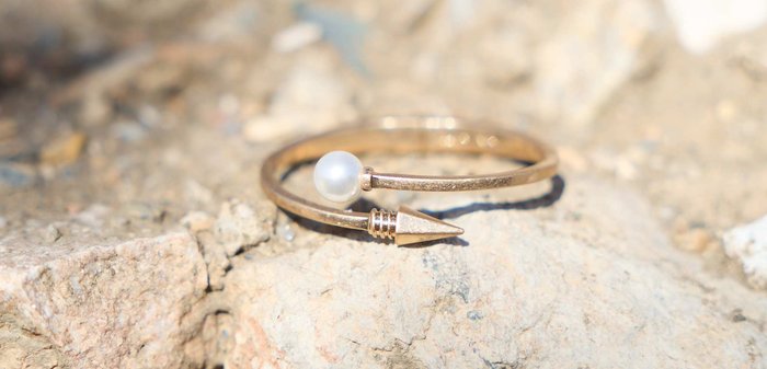 Feiner goldener offener Ring mit einer weißen Perle an einem Ende und einer spitzen Pfeilspitze am anderen, liegend auf einer rauen Steinoberfläche.