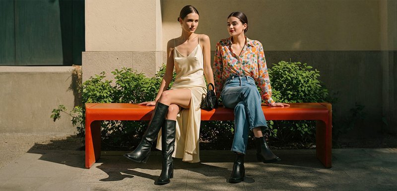 Deux femmes sont assises sur un banc orange. L'une porte une robe en soie crème et des bottes noires jusqu'au genou ; l'autre porte un chemisier fleuri, un jean bleu et des bottes noires à la cheville.