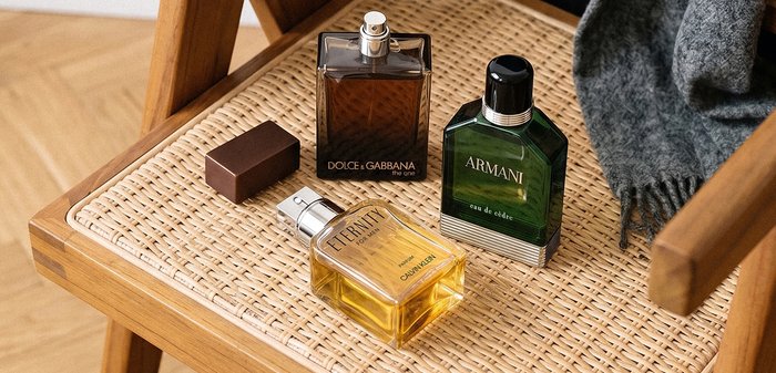 Drie mannen eau de cologne-flessen, waaronder Dolce & Gabbana, Armani en Calvin Klein, op een geweven houten stoelleuning met een grijze stof.