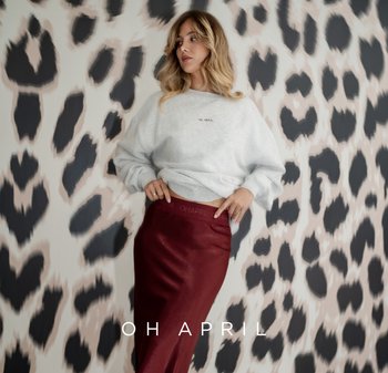 Frau mit blonden Haaren, die einen hellgrauen Pullover und einen hoch taillierten dunkelroten Rock trägt, vor einer großen schwarz-weißen Leopardenmusterwand.